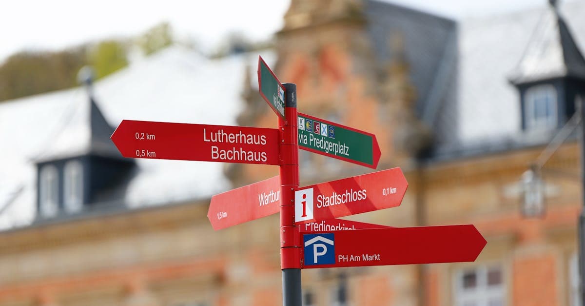 Rotes Wegweiser-Schild mit Sehenswürdigkeiten vor europäischer Architektur zeigt günstiges Reiseziel